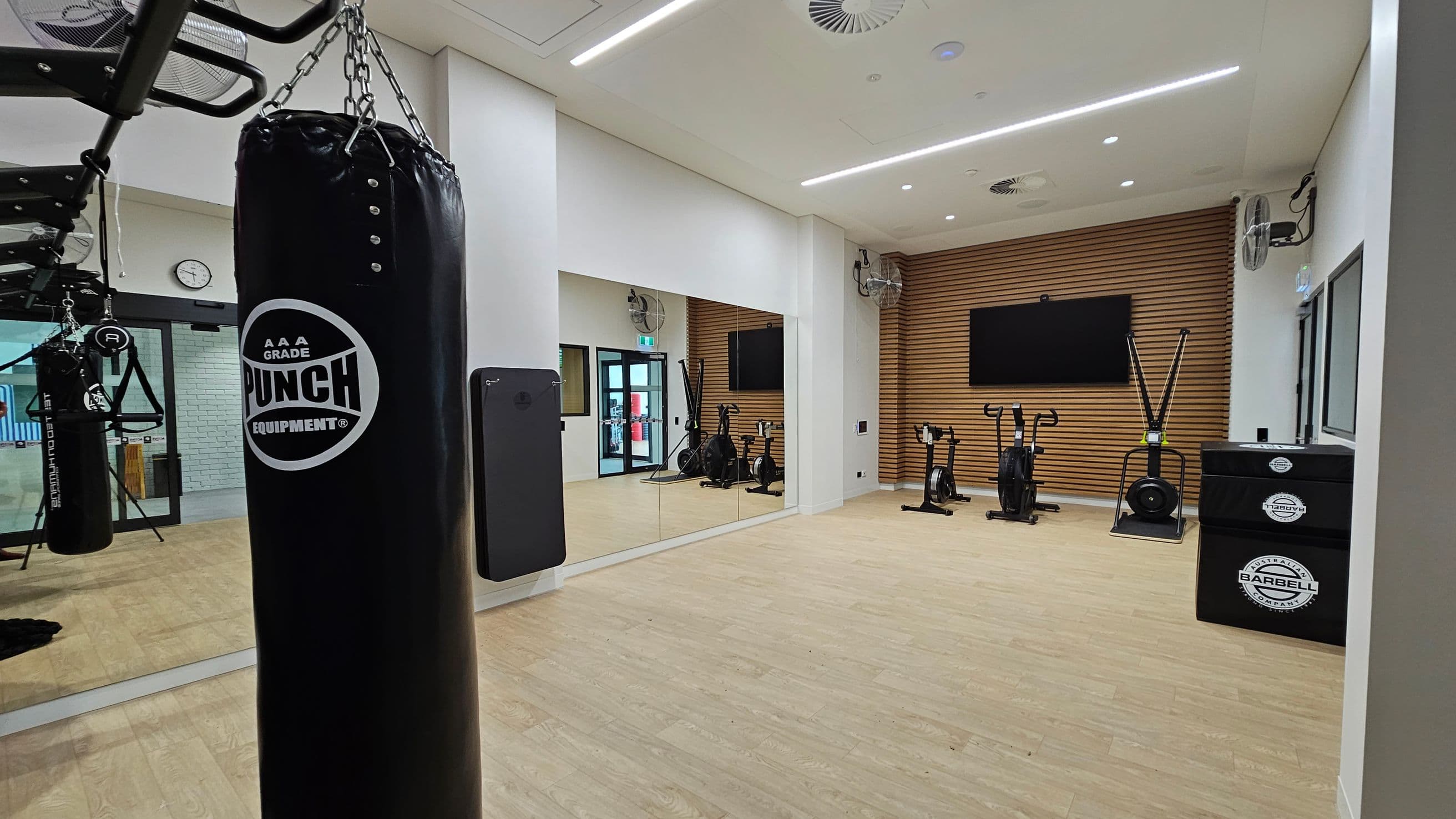 A gym room with a wooden floor and white ceiling. On the left, a black punching bag hangs from the ceiling. A large mirror covers the wall, reflecting the room. In the centre, there are exercise bikes and a rowing machine. The back wall features wooden panelling and a large flat-screen TV. On the right, a black equipment box is visible. Ceiling fans and lights are installed above. A clock and a door are visible on the left wall.