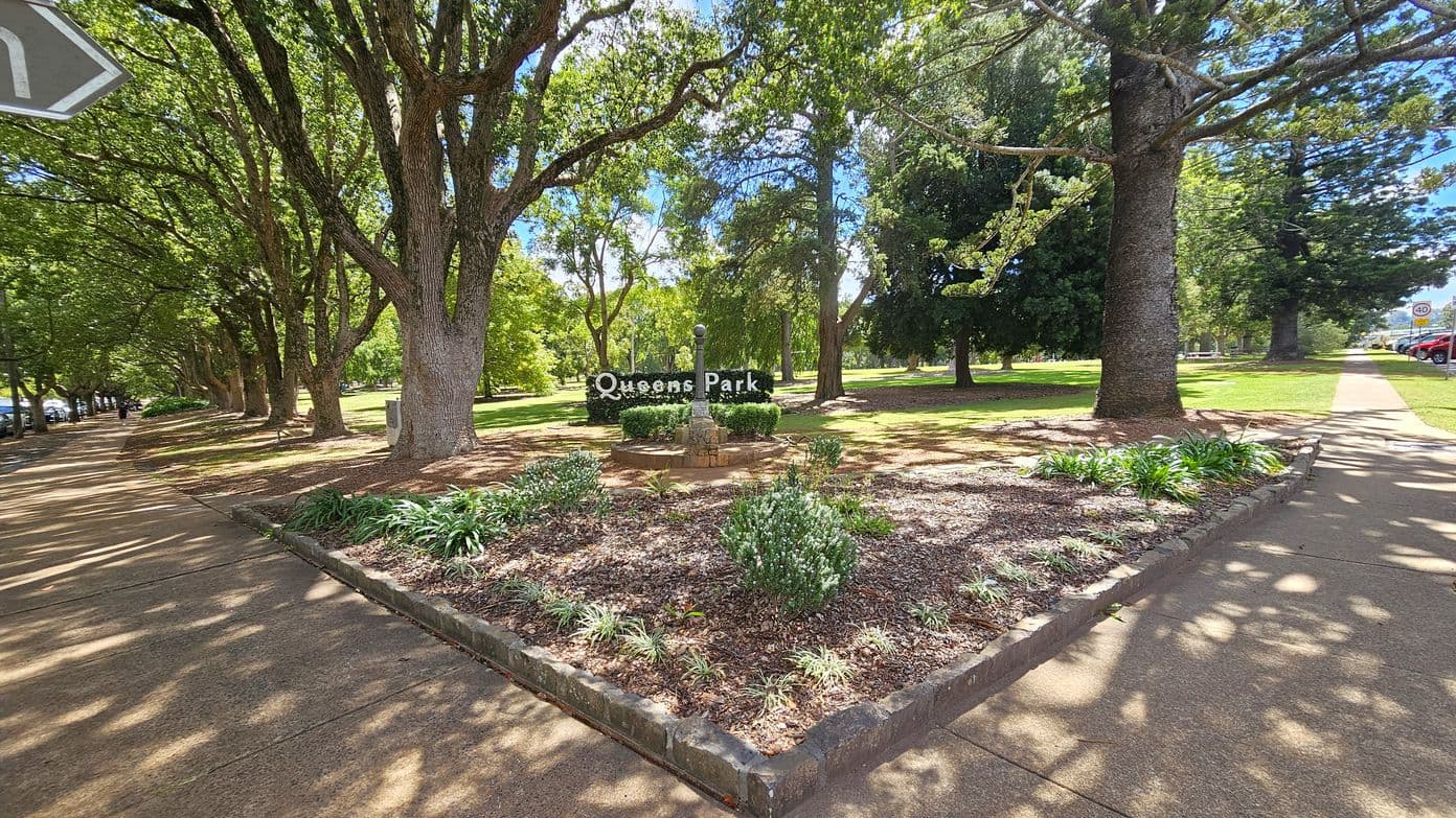 A park scene with a triangular garden bed in the foreground, featuring small shrubs and plants. A stone structure with a pole is centred in the garden bed. Behind it, a sign reads "Queens Park". Tall trees with dense foliage line both sides, casting shadows on the ground. A paved path runs along the left and right sides, with parked cars visible on the right. The area is well-shaded by the trees.