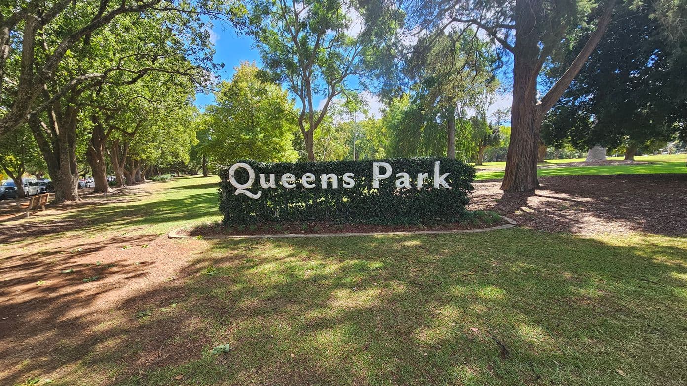 A neatly trimmed hedge with the words "Queens Park" in large white letters is in the centre. Surrounding the hedge are tall trees casting shadows on the grass. To the left, there are benches and parked cars along a path lined with more trees. The ground is covered with grass and patches of dirt, while the background features more greenery and trees.