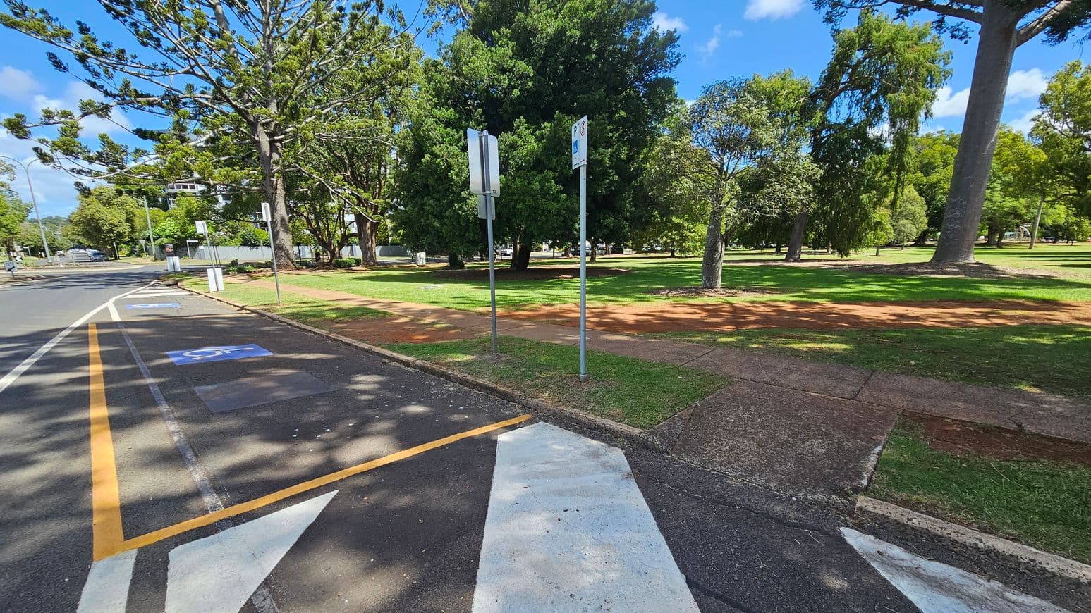 A road with marked parking spaces on the left, including a disabled parking spot. Two signs are visible near the kerb. On the right, a grassy park area with several large trees and a red dirt path. The foreground shows a pedestrian crossing with white stripes. The background includes more trees and a building partially visible through the foliage. Shadows of trees are cast on the road and grass.