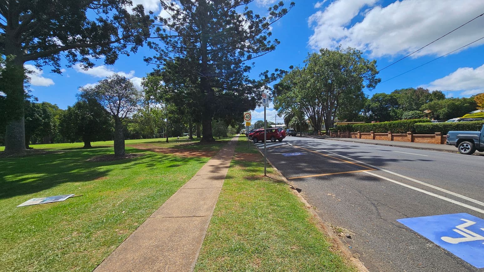 A paved footpath runs through a grassy area on the left, bordered by large trees casting shadows. A road is on the right, with parked cars and a speed limit sign indicating 40. A blue disabled parking symbol is painted on the road. A brick fence with hedges lines the right side, and more trees are visible in the background. A sign lies on the grass near the path.