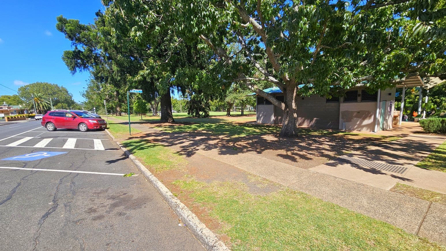 A red car is parked on the left side of a road near a zebra crossing and a disabled parking space. A large tree with dense foliage provides shade on the right, where a small building with a blue roof is partially visible. The ground is a mix of grass and paved pathways. In the background, more trees and a grassy area can be seen, along with a street sign.