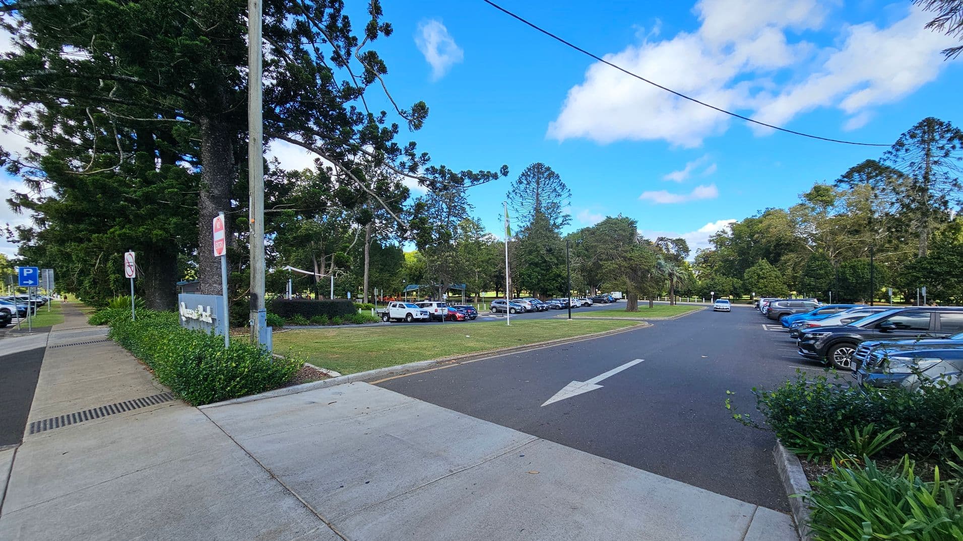 A paved pathway runs along the left side, bordered by lush greenery and large trees. Several signs are visible near the path. On the right, a car park is filled with vehicles, with a clear arrow painted on the road indicating direction. The background features more trees and a grassy area, creating a park-like setting. The sky is bright with scattered clouds.