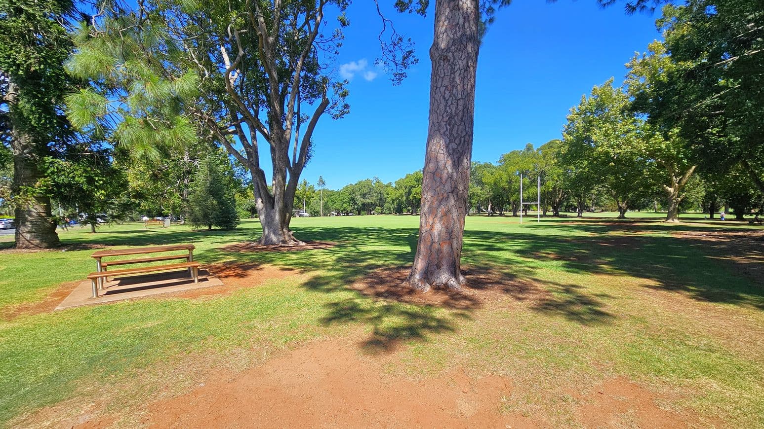 A park scene with a wooden picnic table on the left, surrounded by grass and trees. In the centre, a large tree trunk stands prominently. To the right, more trees and a set of rugby posts are visible. The ground is a mix of grass and patches of bare earth, with shadows cast by the trees. The background features a wide expanse of open grassy field bordered by trees.
