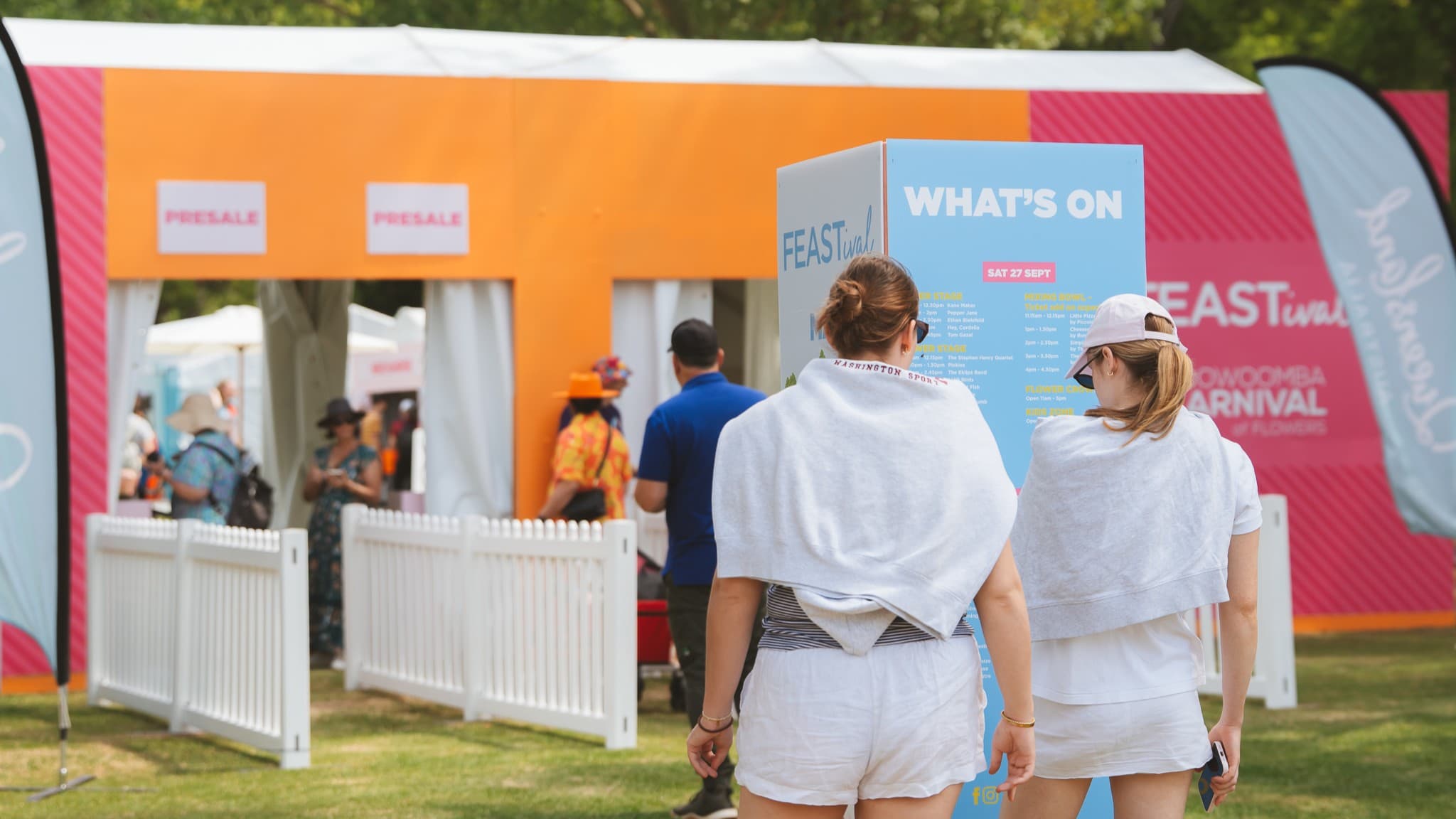 Two people in white clothing and caps stand in the foreground, facing a blue sign with "WHAT'S ON" written on it. They are near a white picket fence. In the background, there is an orange and pink tent with people walking around. The ground is grassy, and there are trees visible in the distance.