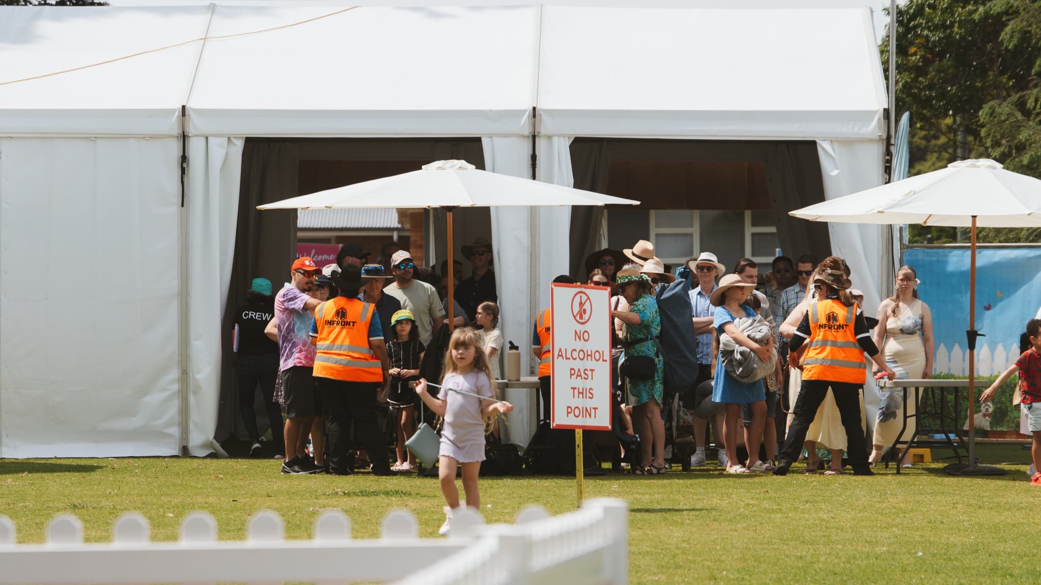 A large white tent with people gathered at the entrance. Two individuals in orange high-visibility vests are managing the crowd. A sign reads "No Alcohol Past This Point." Two white umbrellas provide shade. A child in the foreground walks on the grass, which is bordered by a white picket fence. The background includes a blue banner with a fence design and some trees.