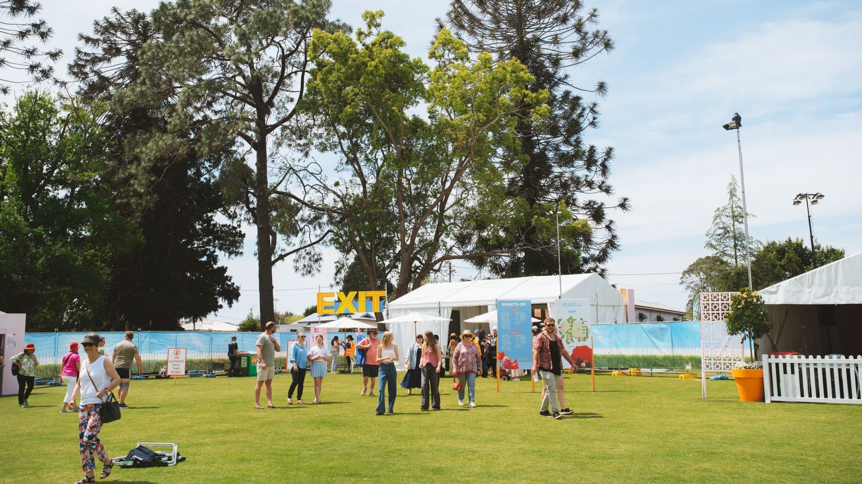 A grassy outdoor area with people walking around. On the left, a few individuals are near a pink structure. In the centre, a large yellow "EXIT" sign is visible above a white tent. Several people are gathered near informational boards. On the right, a white picket fence and a tall light pole are present. Tall trees are in the background, providing shade.