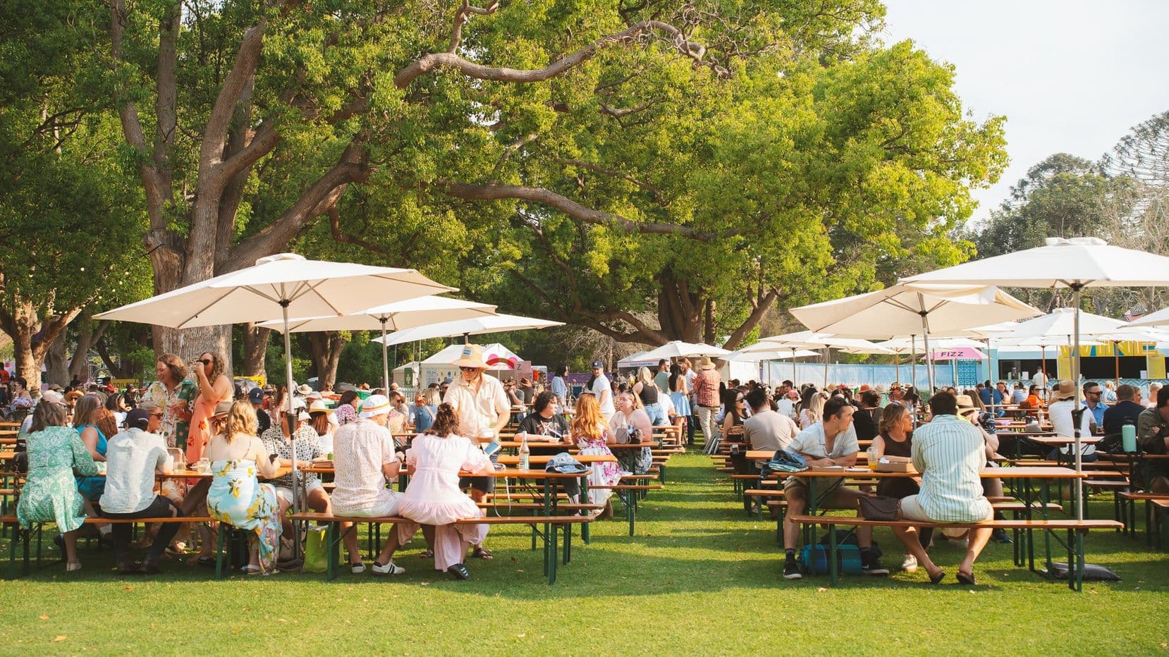 People are seated at long wooden picnic tables under large white umbrellas on a grassy area. The tables are arranged in rows, with many individuals enjoying food and drinks. In the background, there are large trees providing shade, and several food stalls or trucks are visible. The scene is lively, with people dressed in casual summer attire, suggesting a relaxed outdoor event or festival.