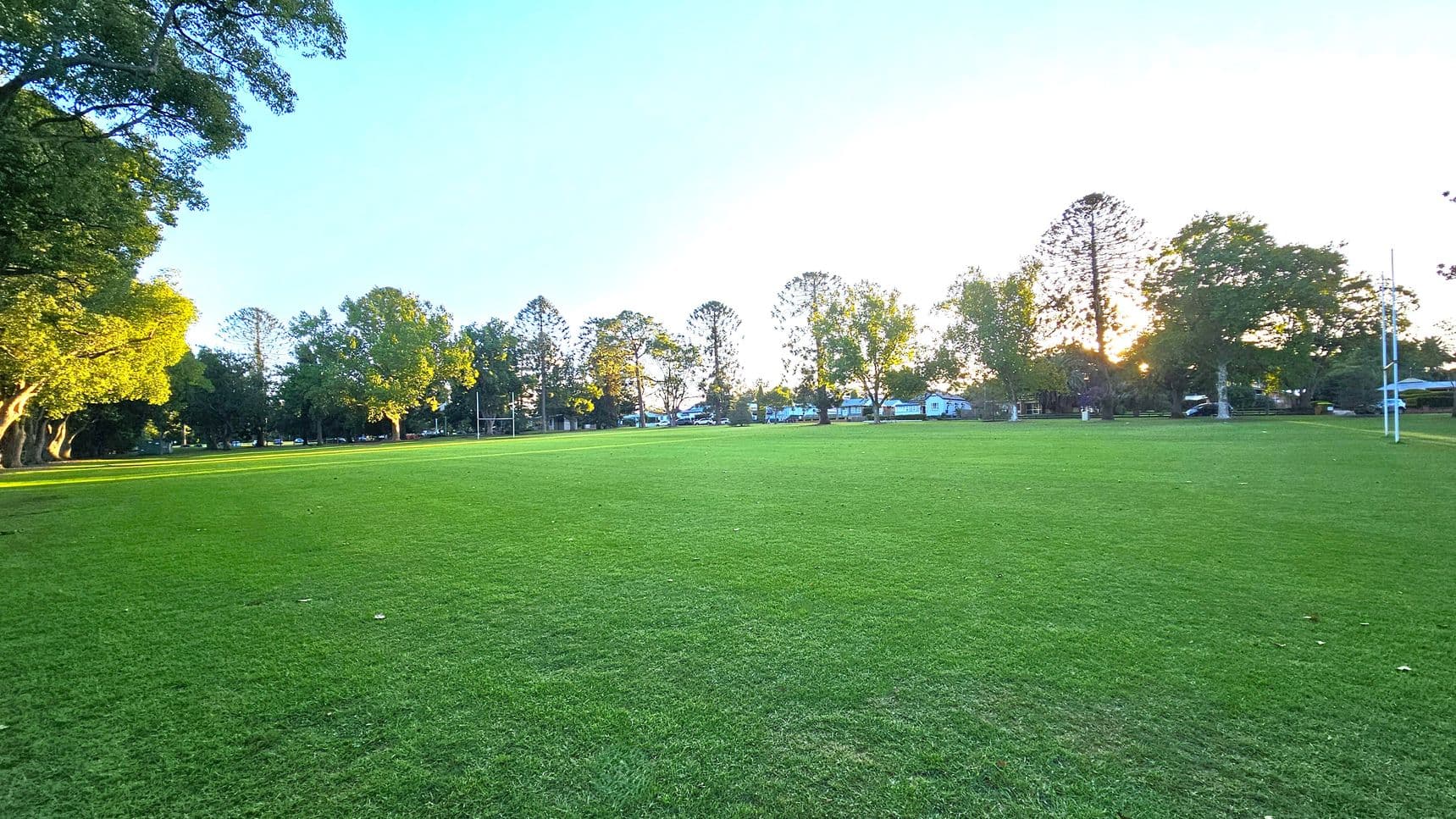 A wide, open grassy field with rugby posts on the right. Trees line the background, with sunlight filtering through, casting long shadows. On the left, more trees create a shaded area. The grass is lush and green, and the scene is peaceful and expansive.