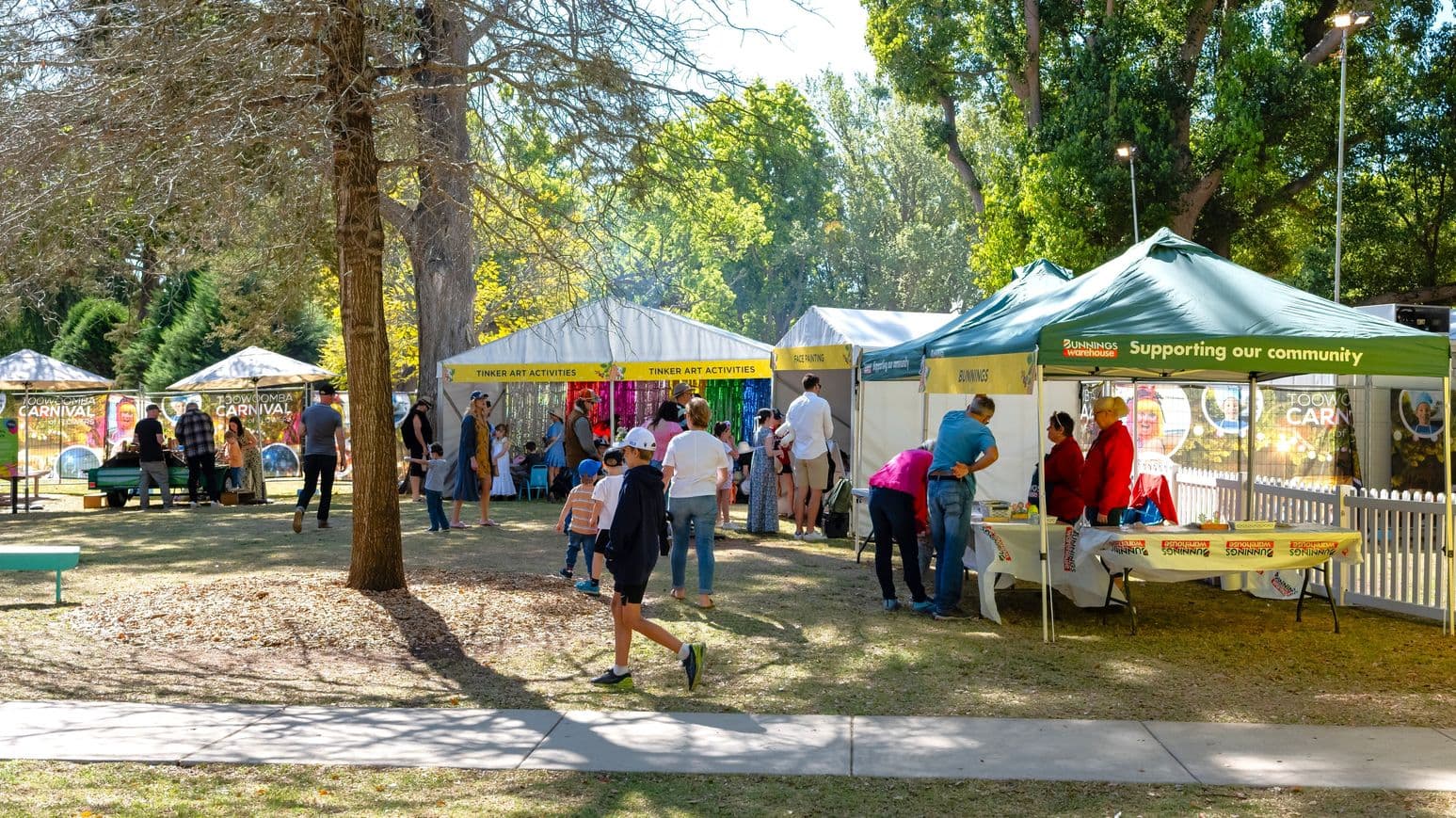 On the left, a tree stands with people gathered around tables under umbrellas. In the centre, several tents with signs like "Tinker Art Activities" and "Face Painting" are set up, with people, including children, participating in activities. On the right, a green tent with "Bunnings Warehouse" branding has people interacting at a table. The background is filled with lush green trees, and the ground is covered with grass and a few fallen leaves.