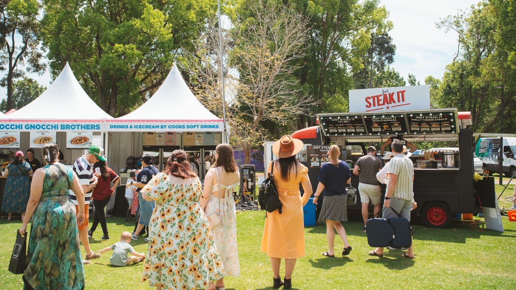 A lively outdoor food festival scene with people gathered around two food stalls. On the left, a white tent with "Gnocchi Gnocchi Brothers" signage offers drinks and ice cream. On the right, a black food truck with a "Steak It" sign serves customers. People are dressed in casual summer attire, with one woman in a yellow dress and hat. The background features tall green trees, and the ground is covered with grass.