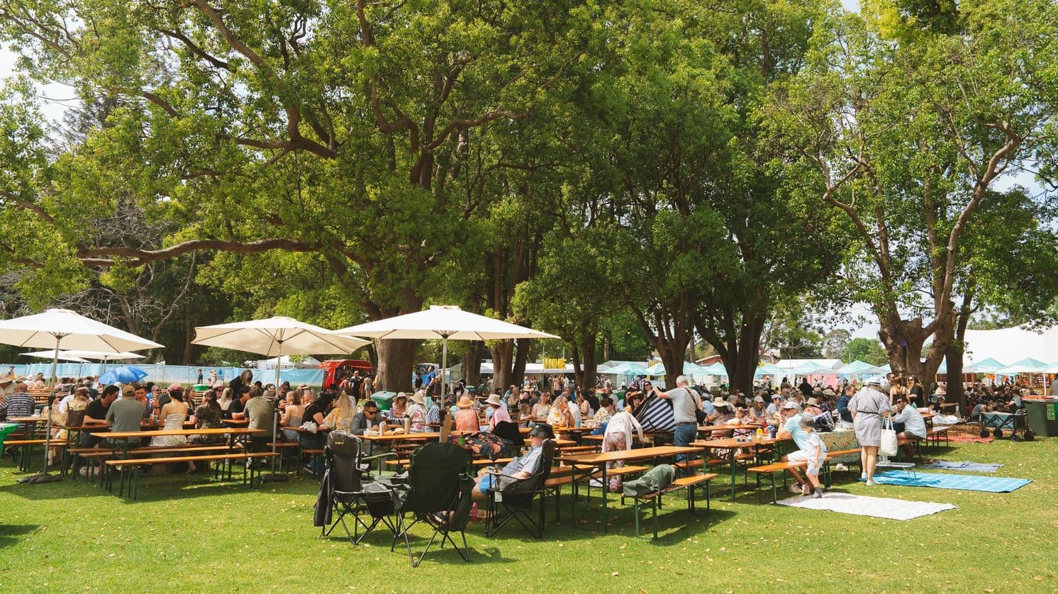 A lively outdoor scene with people gathered around wooden picnic tables under large white umbrellas. The tables are set on a grassy area, surrounded by tall, leafy trees providing shade. In the background, there are more people and stalls with colourful canopies, suggesting a market or festival atmosphere. Some individuals are seated on blankets on the grass, enjoying the day. The setting is bright and bustling with activity.