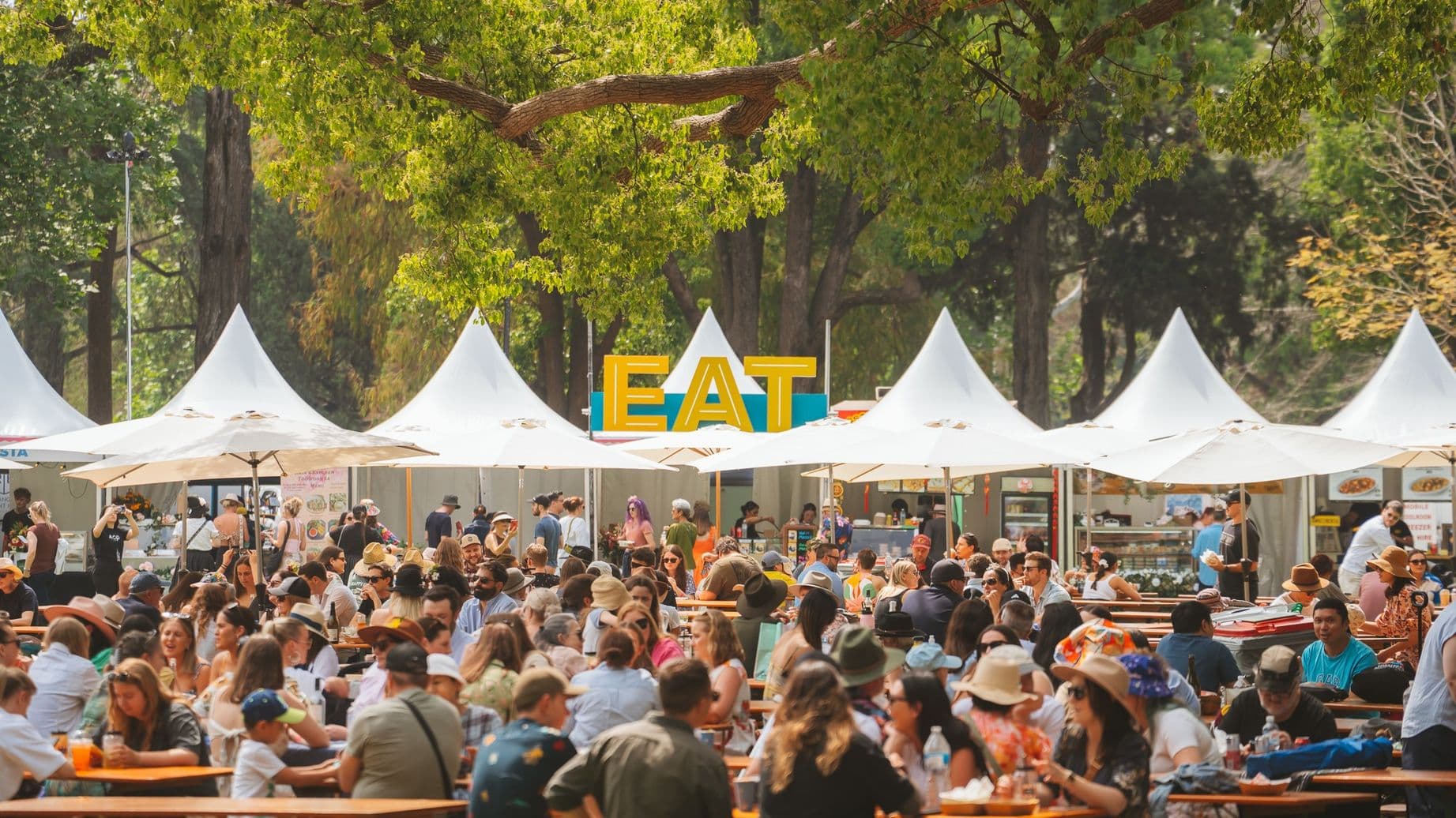 A bustling outdoor food festival with numerous people seated at long wooden tables, enjoying meals. White tents with pointed tops line the background, with a large, colourful "EAT" sign prominently displayed. The area is shaded by large trees with green foliage. Many attendees wear hats and sunglasses, suggesting a sunny day.