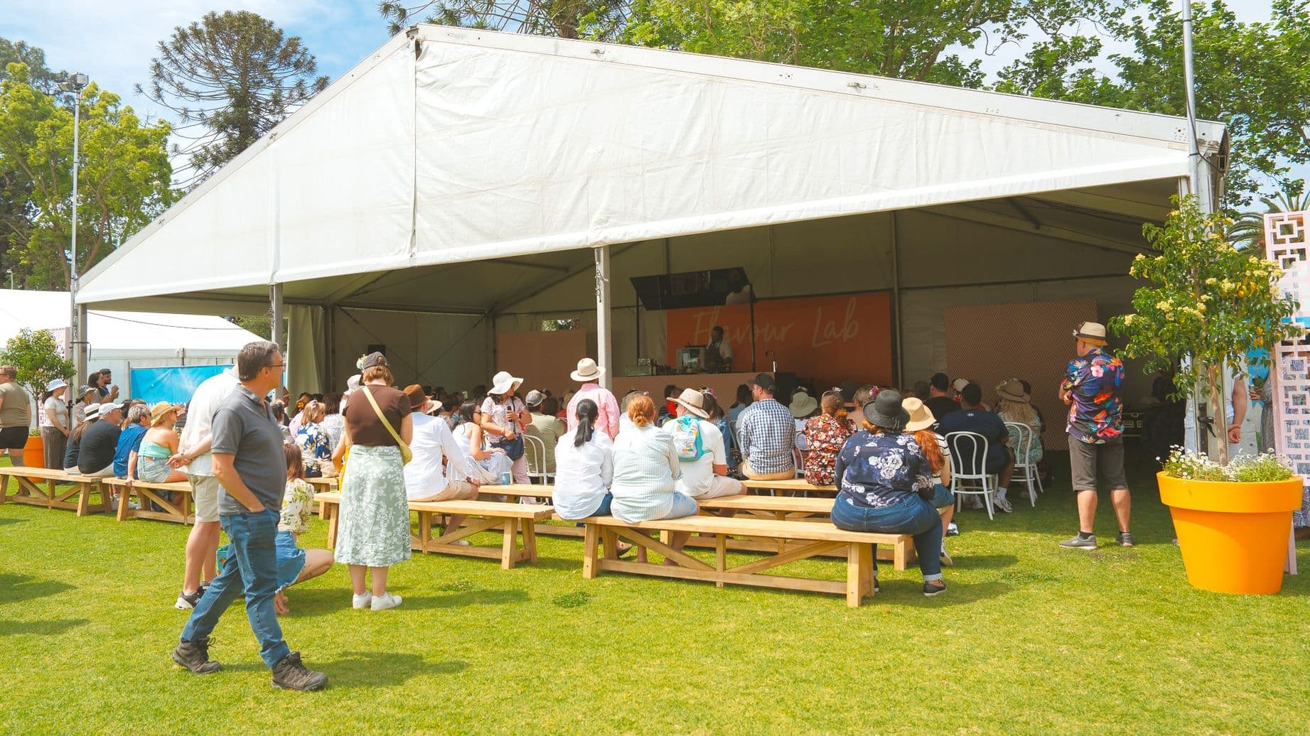A large white tent is set up on a grassy area. Inside, people are seated on wooden benches, facing a stage where a presentation appears to be taking place. Many attendees are wearing hats, and some are standing. To the right, there is a large orange planter with a tree. In the background, tall trees are visible, and the tent is open on one side, allowing a view of the audience and the speaker.