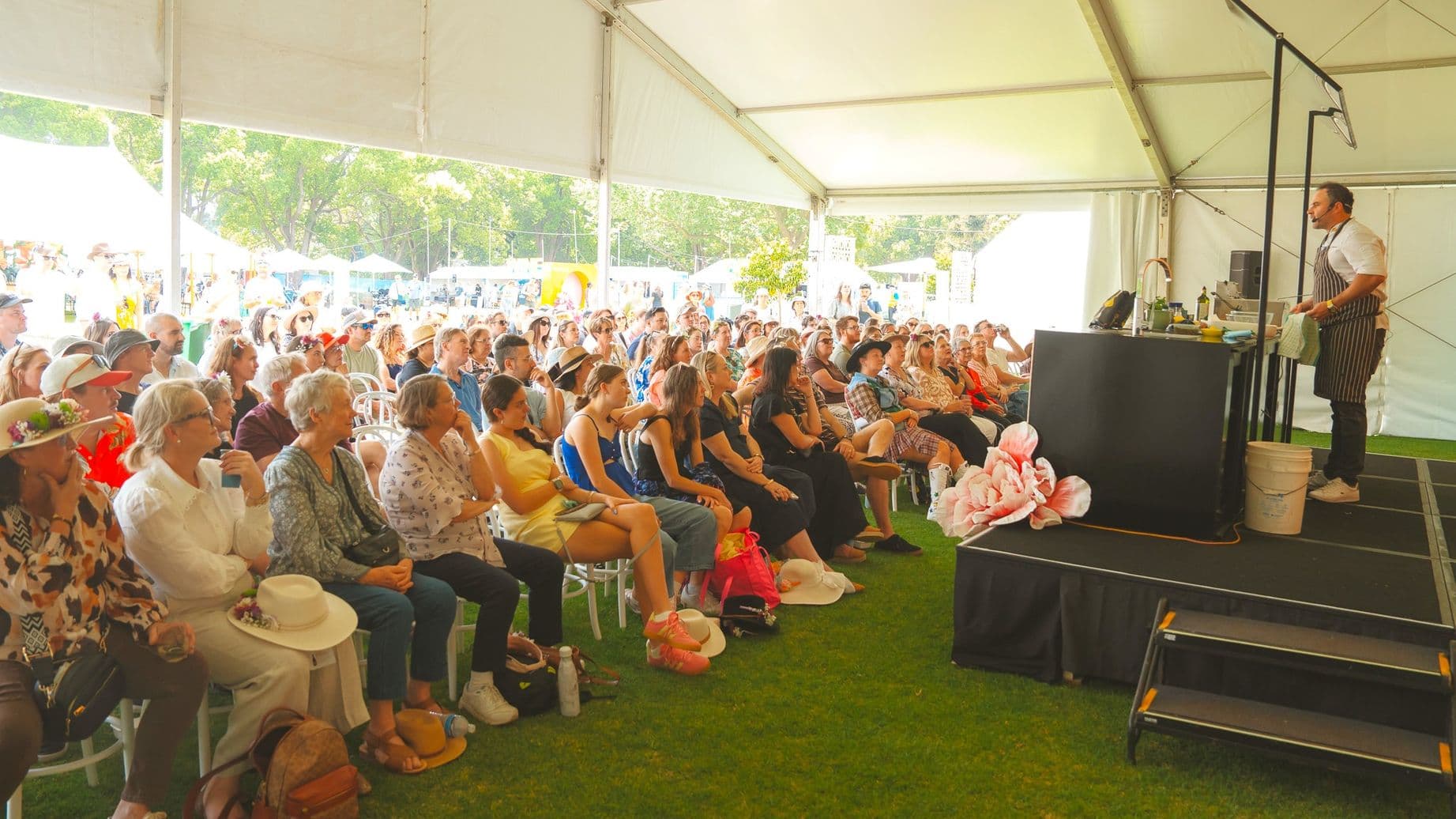 A large group of people is seated under a white tent, attentively watching a cooking demonstration. The audience, consisting of various ages, is seated on white chairs on a grassy floor. On the right, a person in an apron stands behind a counter with cooking equipment. The background shows a bright outdoor area with trees and additional tents. The ceiling of the tent is white and supported by metal frames.