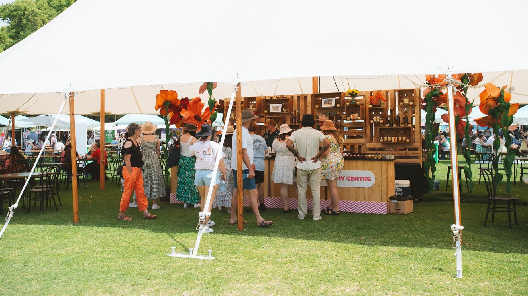 Under a large white tent, people are gathered around a wooden stall displaying various products. The stall is decorated with large orange flowers. The ground is covered with grass, and there are tables and chairs scattered around, with more people seated in the background. The tent is supported by wooden poles, and the atmosphere suggests an outdoor market or festival setting.
