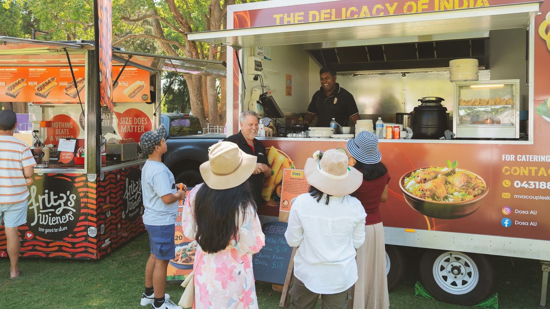 A food truck scene with two adjacent stalls. On the left, a stall with orange signage advertises hot dogs, featuring phrases like "Nothing Beats" and "Size Does Matter." A man in a striped shirt stands in front. On the right, an Indian food truck displays images of dishes and contact information. A man inside serves customers. Several people, including women in hats, are gathered in front, looking at the menu. The setting is outdoors with trees in the background.
