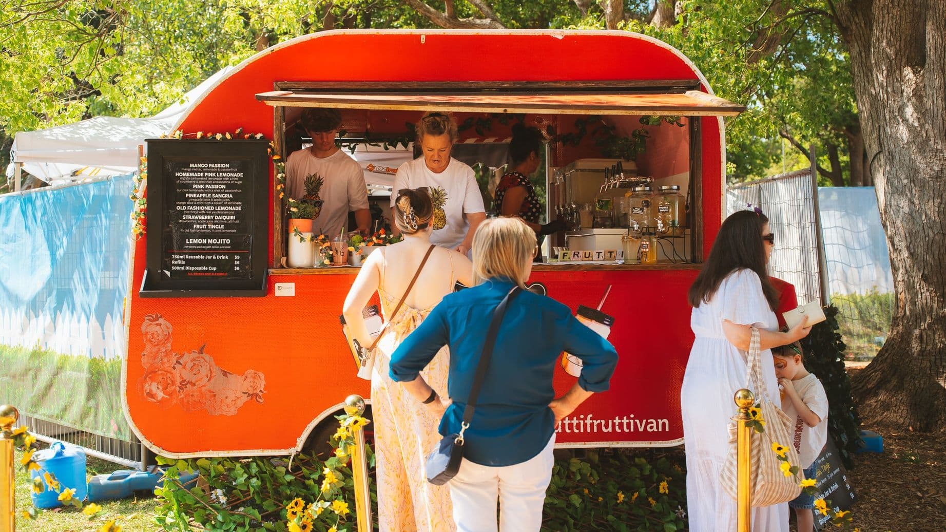 A vibrant red food truck is parked outdoors, surrounded by greenery. On the left, a menu board lists various drinks. In front of the truck, several people are gathered, including a woman in a blue shirt and another in a white dress with a child. Inside the truck, staff members are preparing orders. The ground is grassy, and there are decorative sunflowers around the area. A large tree is visible on the right, providing shade.