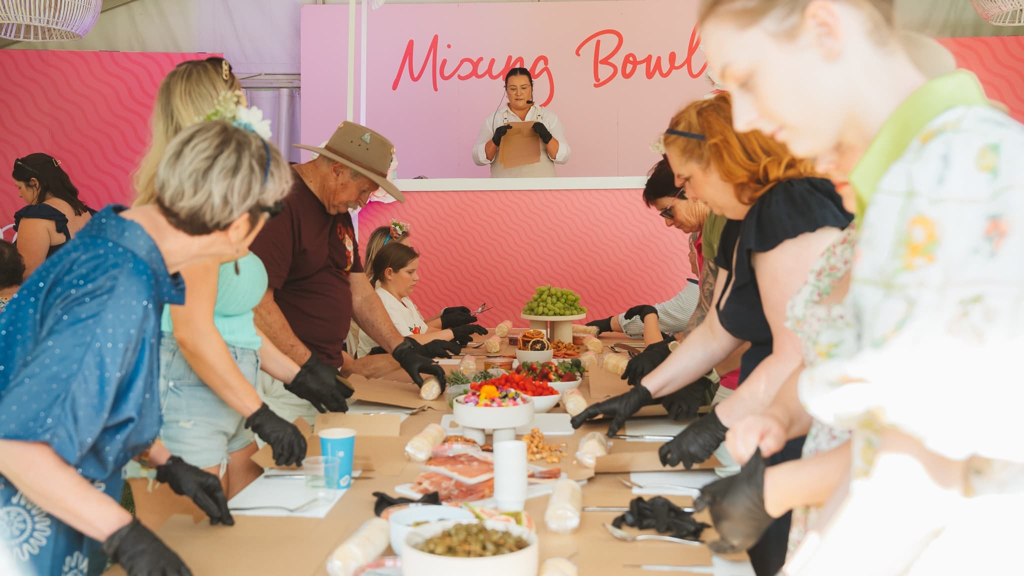 A group of people are gathered around a long table, each wearing black gloves and focused on preparing food items. The table is filled with various ingredients, including grapes, strawberries, and other foods. In the background, a person stands behind a counter with a sign that reads "Mixing Bowl" in pink letters. The setting appears to be a cooking class or workshop, with participants engaged in a hands-on activity. The ceiling features a white, woven light fixture.