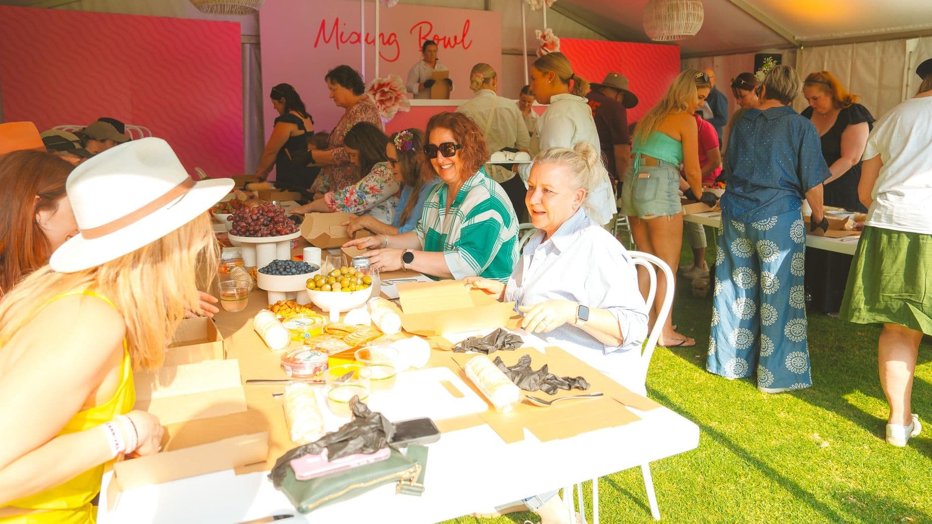 A lively indoor gathering with people seated and standing around tables. On the left, a woman in a white hat is engaged in conversation. In the centre, two women are smiling, with various foods like grapes, blueberries, and olives on the table. The background features a pink wall with the words "Missing Bowl" and more people interacting. The floor is covered with green grass, and the ceiling has a white, tent-like structure with hanging lights.