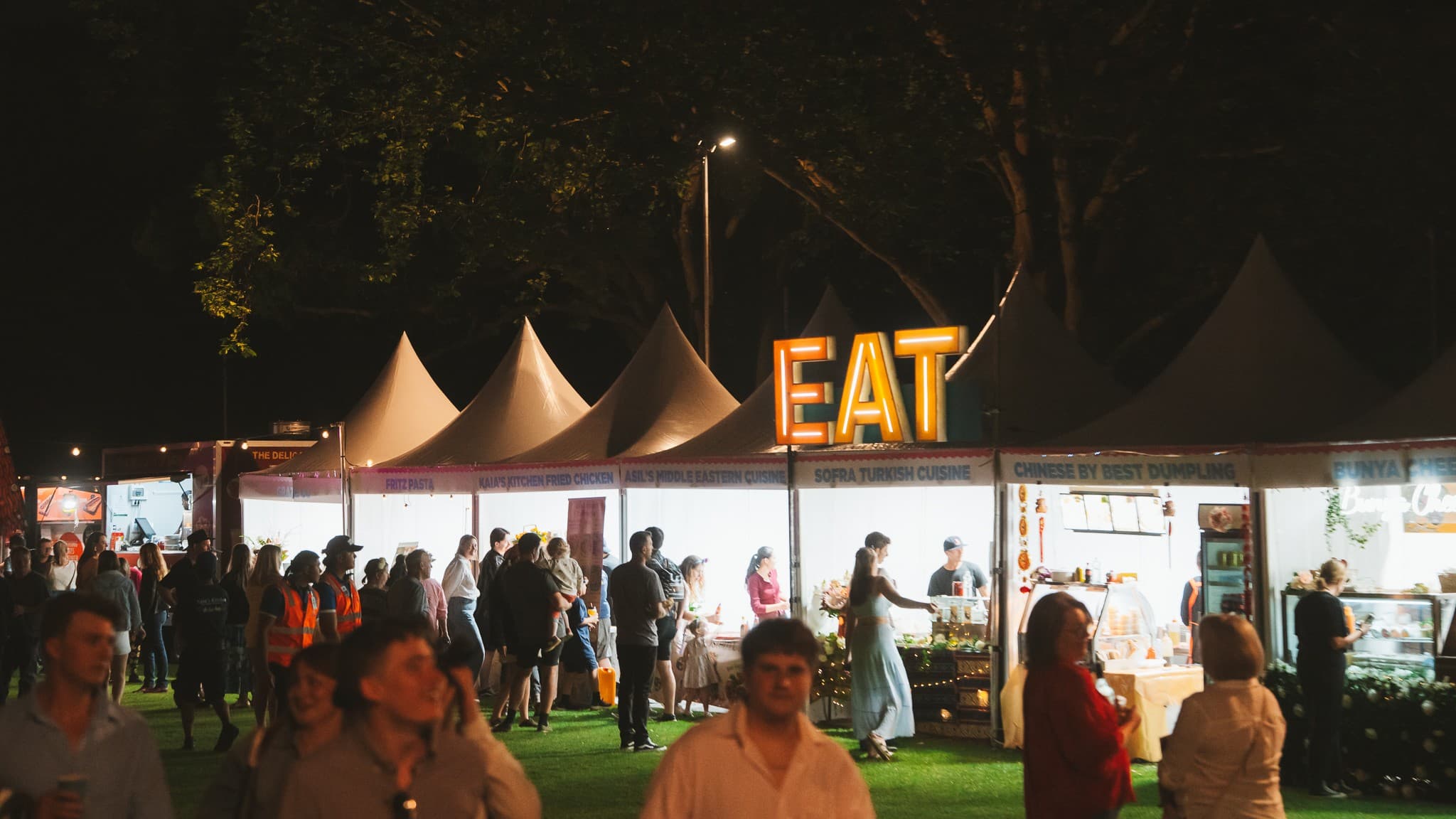A night-time food festival scene with several white tents lined up in the background. Each tent is brightly lit, showcasing various food stalls. A large illuminated sign reading "EAT" is prominently displayed above one of the stalls. People are gathered around the stalls, some queuing and others walking past. The ground is covered with grass, and there are trees visible in the background. The atmosphere is lively with a mix of people enjoying the event.
