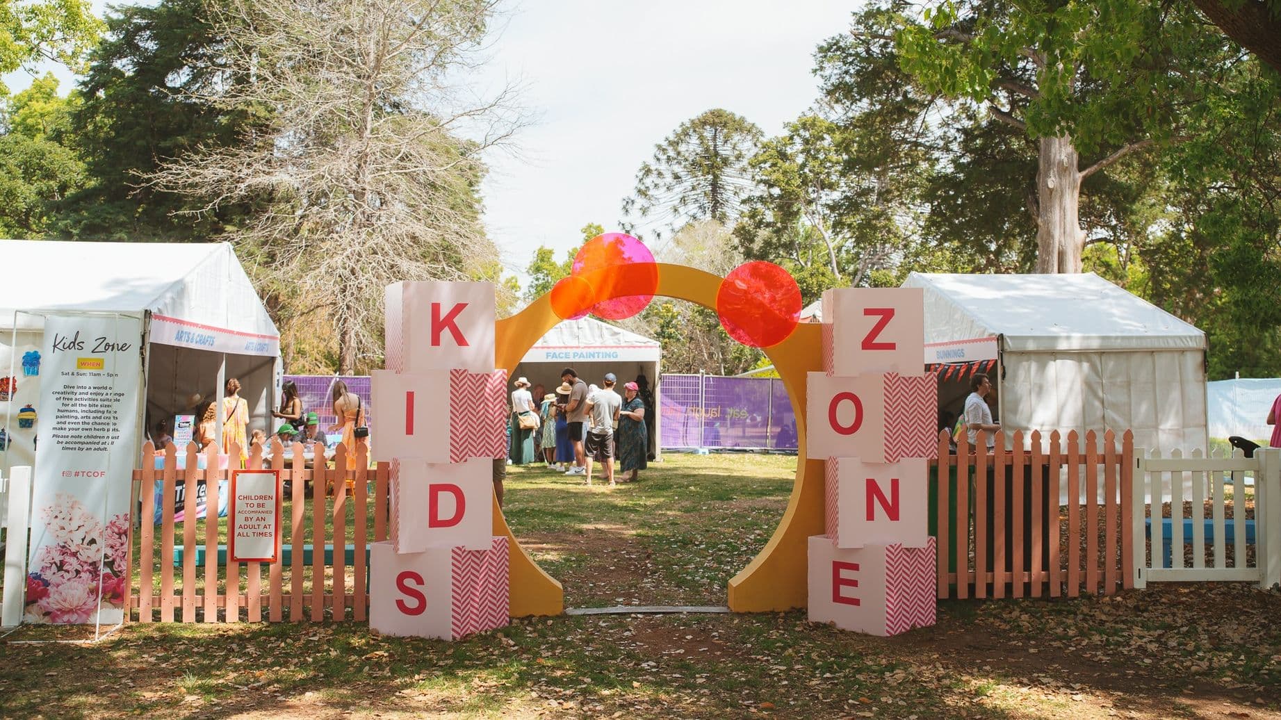 A colourful entrance to a "Kids Zone" with large blocks spelling out "KIDS" on the left and "ZONE" on the right. The entrance is framed by an arch with red and orange circular decorations. Behind the entrance, people are gathered under white tents, with trees and greenery in the background. A wooden picket fence surrounds the area, and a sign on the fence reads, "CHILDREN TO BE SUPERVISED BY AN ADULT AT ALL TIMES." The ground is covered with grass and fallen leaves.