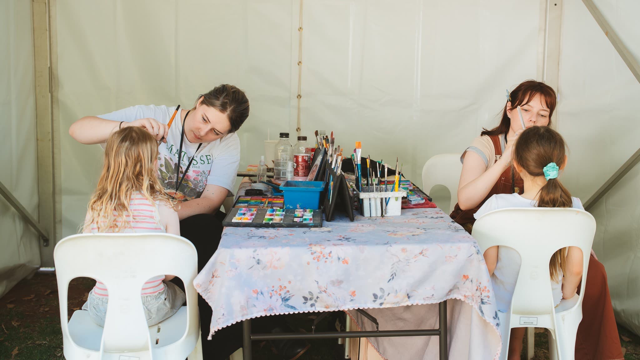 Two women are face painting two children seated on white plastic chairs. The table between them is covered with a floral cloth and holds various paints, brushes, and containers. The background is a light-coloured tent wall. The floor is grassy, and the ceiling is not visible.