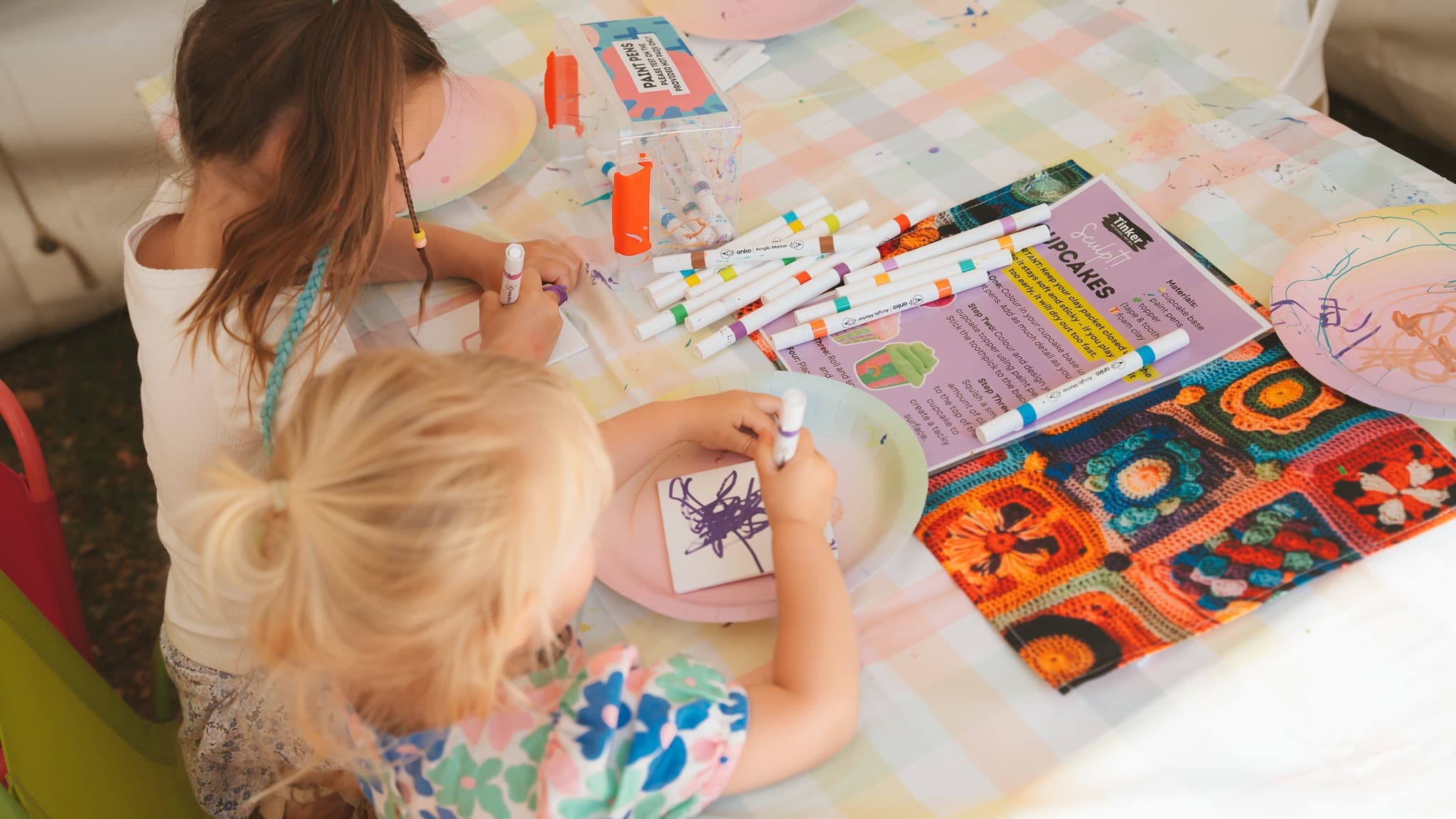 Two children are seated at a table covered with a pastel-checkered tablecloth. They are drawing on paper plates with colourful markers. A variety of markers are scattered on the table, along with a clear box labelled "PAINT PENS". A colourful crochet-patterned cloth is partially visible on the right. A printed sheet with instructions or information is also on the table. The children are focused on their artwork, with one child having a blue braid in their hair.