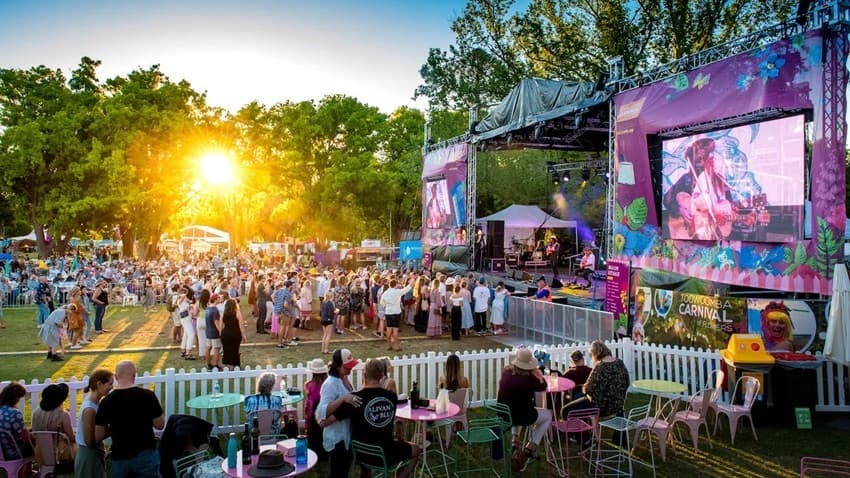A lively outdoor festival scene with a large stage on the right, featuring colourful floral decorations and two large screens displaying performers. In the middle, a crowd gathers in front of the stage, enjoying the performance. On the left, the sun sets behind tall trees, casting a warm glow over the event. In the foreground, people sit at tables with pastel-coloured chairs, enclosed by a white picket fence. The atmosphere is vibrant and bustling.