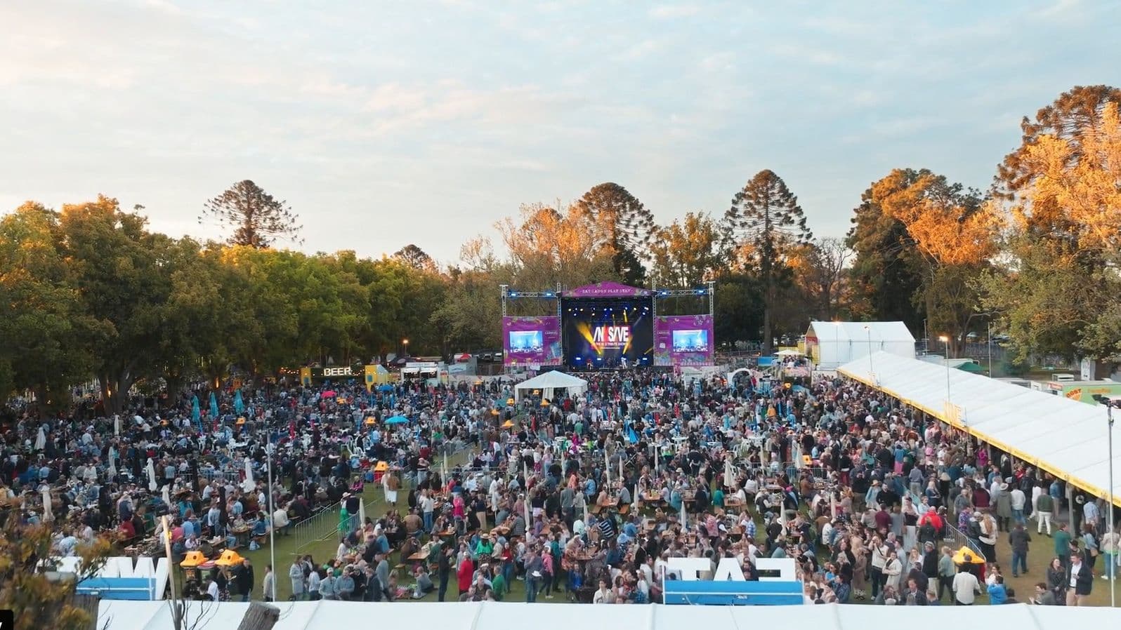 A large outdoor festival with a crowd gathered in front of a stage displaying "MASSIVE" on the screen. The stage is flanked by two smaller screens. On the left, there are trees and a sign for "BEER" among the crowd. On the right, a long white tent stretches along the side. The background features tall trees with autumn foliage. The ground is grassy, and the area is bustling with people.
