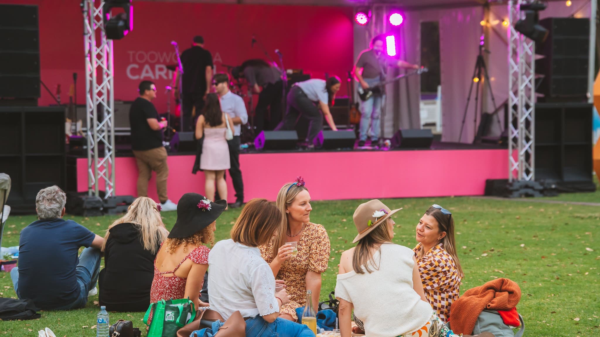 A group of people sit on the grass in the foreground, some wearing hats and floral attire, engaged in conversation. In the background, a stage with pink lighting and a band setting up is visible. The stage is framed by metal trusses and large speakers. The grass is dotted with a few items like bags and a water bottle. The atmosphere suggests a casual outdoor event or festival.