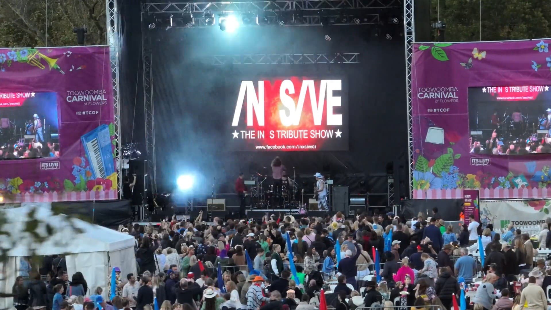 A large outdoor stage with a crowd gathered in front. The stage features a big screen displaying "INXSIVE: The INXS Tribute Show" with bright lights illuminating the area. On either side of the stage are large banners for the Toowoomba Carnival of Flowers, decorated with floral and musical motifs. The audience is diverse, with many people holding colourful inflatable objects. The atmosphere is lively, with people enjoying the performance.