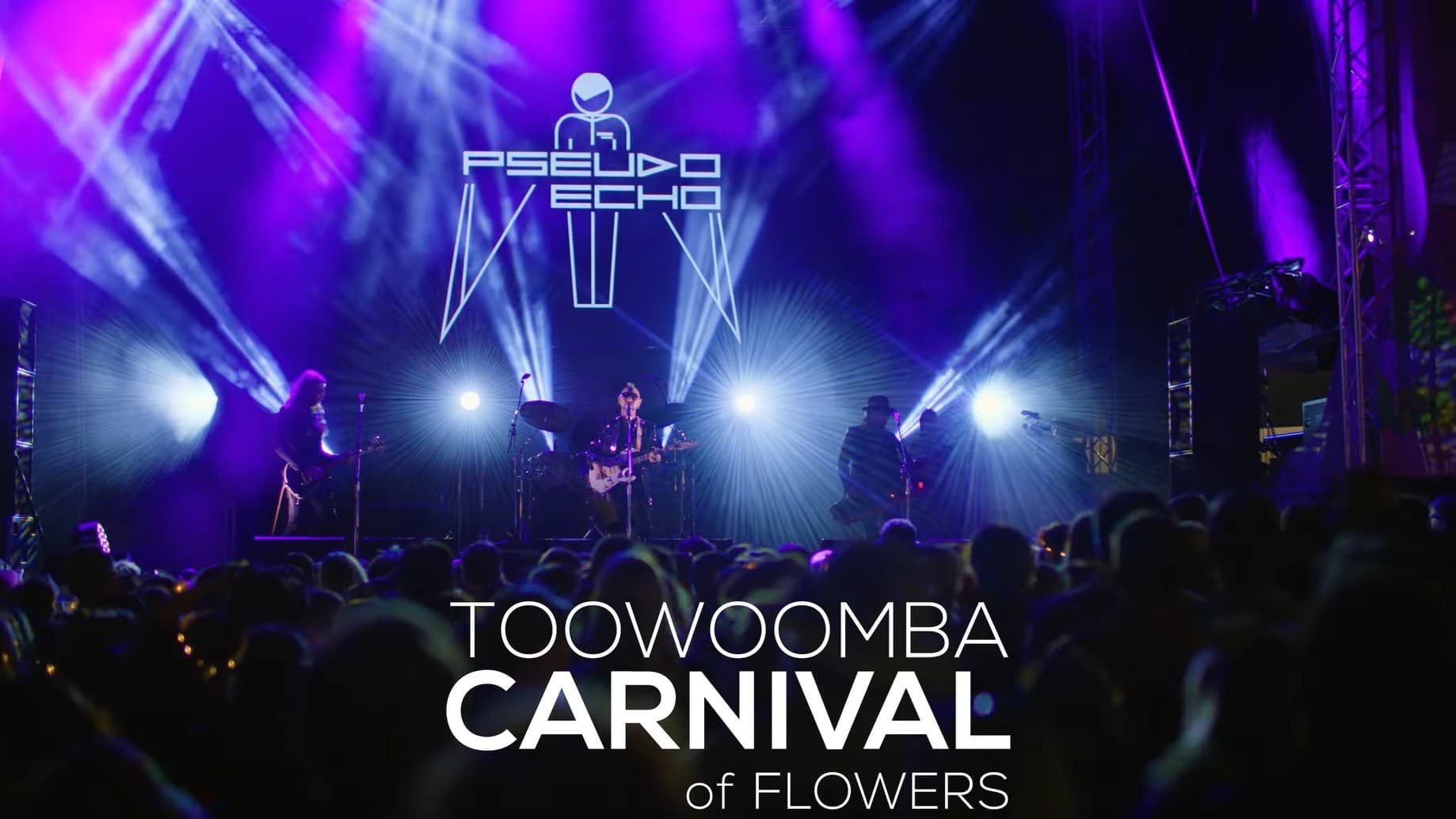 A concert stage with a band performing under vibrant purple and blue lighting. The band is silhouetted against bright spotlights, with a logo reading "PSEUDO ECHO" above them. The audience is visible in the foreground, with the words "TOOWOOMBA CARNIVAL of FLOWERS" prominently displayed at the bottom. The stage is flanked by tall speaker towers, and beams of light create dynamic patterns across the scene.