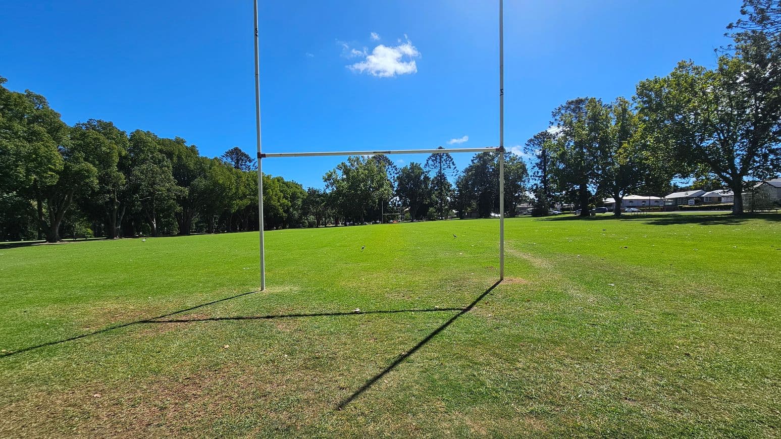 A rugby goalpost stands in the centre of a grassy field. The foreground shows a mix of green and brown grass, with the shadow of the goalpost extending towards the viewer. In the background, a line of trees borders the field, with some houses visible to the right. The sky is clear and blue.