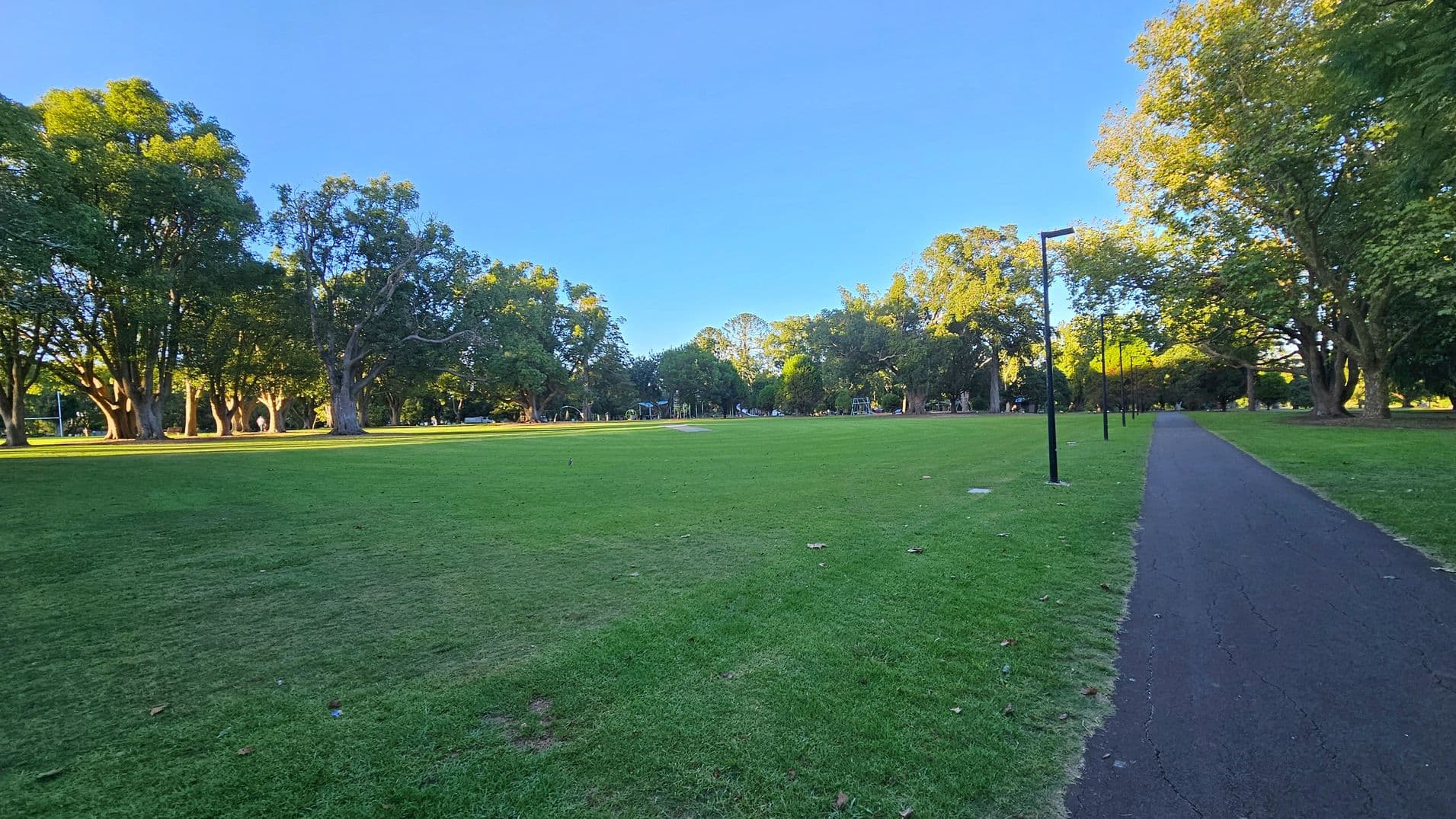 A park with a large grassy area in the centre, surrounded by tall trees. On the right, a paved path runs parallel to a row of black lamp posts. The trees on both sides have dense foliage, casting shadows on the grass. The ground is covered with well-maintained grass, and the sky is clear.