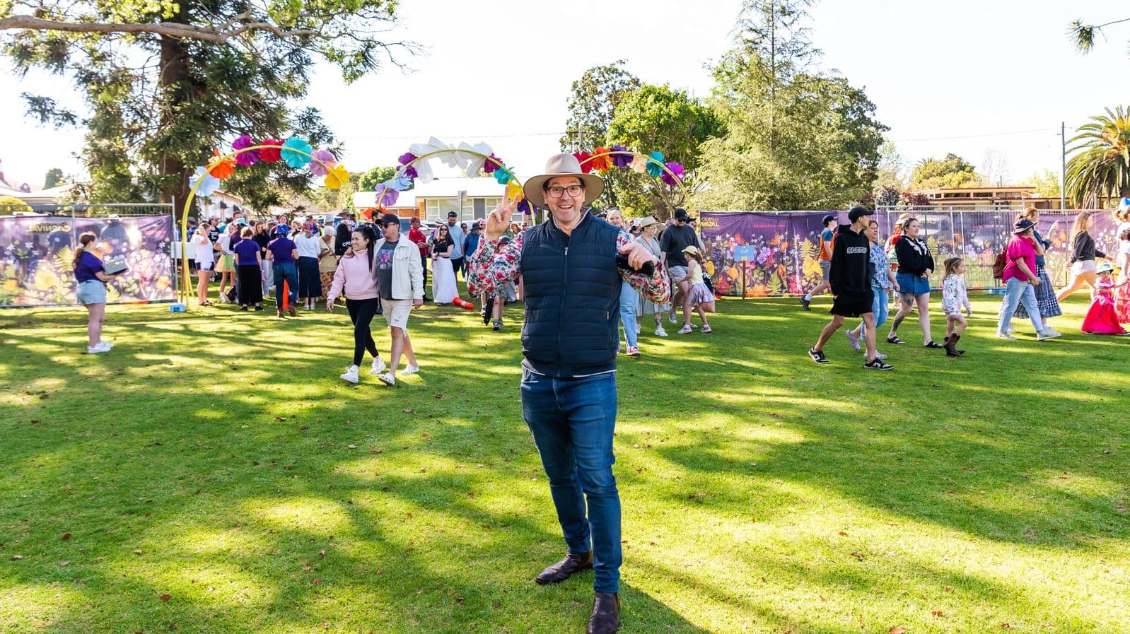 A man wearing a hat, floral shirt, and dark vest stands on a grassy area, gesturing with both hands. Behind him, people walk and gather under a colourful arch made of large, vibrant flowers. To the left, a person in shorts holds a clipboard. In the background, trees and a fence with floral banners are visible. The scene is lively, with people of various ages enjoying the outdoor setting.