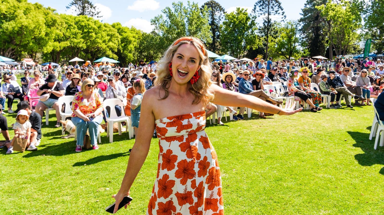 A woman in a strapless dress with an orange floral pattern stands on a grassy area, smiling and gesturing with one arm extended. She holds a phone in her other hand. Behind her, a large crowd of people is seated on white plastic chairs, with some wearing hats and sunglasses. The background features green trees and several market-style tents. The scene suggests an outdoor event or festival on a sunny day.