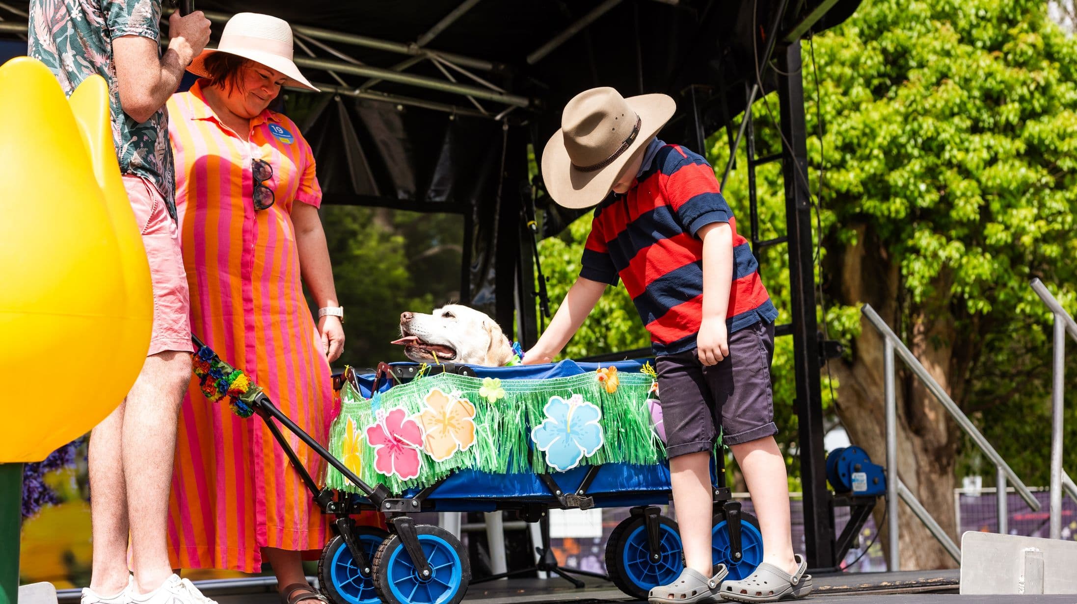 A woman in a pink and orange striped dress and a wide-brimmed hat stands on the left, smiling at a dog in a decorated cart. The cart is adorned with green fringe and colourful flower cutouts. A child in a red and black striped shirt and a wide-brimmed hat is on the right, gently touching the dog. The background features a stage with a black canopy and green trees. The floor is a dark stage surface, and there are metal steps leading down.