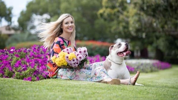 A woman with long blonde hair sits on a grassy lawn, holding a bouquet of colourful flowers. She is wearing a vibrant, patterned dress. Beside her, a large dog with a white and brown coat sits with its tongue out. In the background, there are lush green trees and a bed of bright purple flowers. The scene is outdoors, with a natural and serene setting.