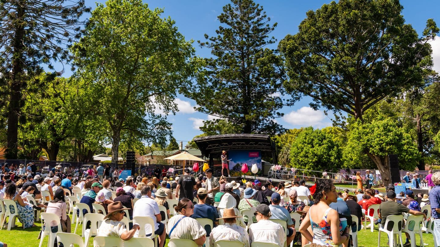 A large outdoor gathering with people seated on white plastic chairs on a grassy area. In the centre, a stage is set up with a person speaking into a microphone. The background features tall trees with dense green foliage. The audience is diverse, wearing casual clothing and hats, suggesting a sunny day. The stage is decorated with colourful elements, and there are additional people standing around the perimeter.