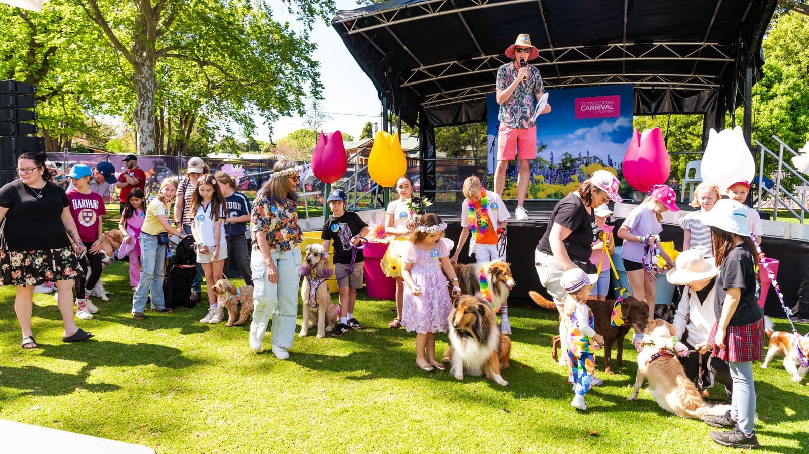 A lively outdoor event with people and dogs gathered on a grassy area. On the left, a group of people, including children, are interacting with their dogs. In the centre, a man in a hat and colourful attire stands on a stage with a microphone. Large, colourful tulip decorations are visible on the stage. On the right, more people, including children, are engaging with their dogs. Trees and a fence are in the background, providing a park-like setting.
