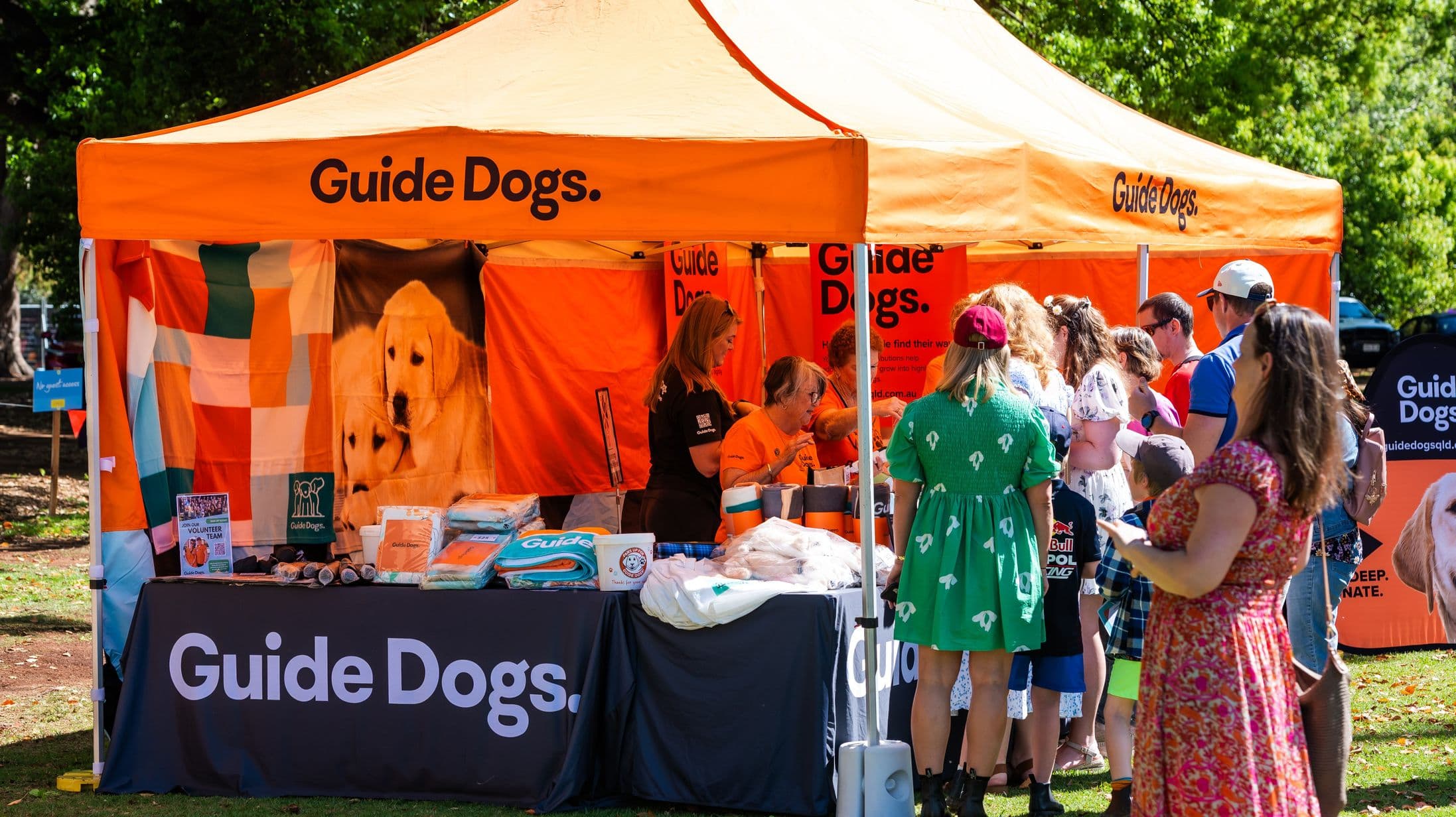 An outdoor event features an orange tent with "Guide Dogs" written on it. Inside, volunteers are interacting with visitors. The table in front displays various items, including brochures and merchandise. A large banner with images of dogs is visible. People are gathered around, some wearing colourful clothing. The background includes trees and a grassy area, suggesting a park setting.