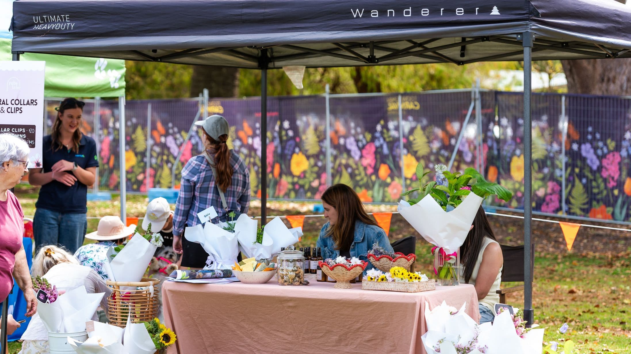 A market stall under a black canopy with "Wanderer" branding. The table is covered with a beige cloth and displays various items, including wrapped flowers, jars, and small baskets. Two women are seated behind the table, one looking down. On the left, a sign advertises floral collars and clips. Several people are browsing, and a colourful floral mural is visible in the background. The ground is grassy with scattered leaves.
