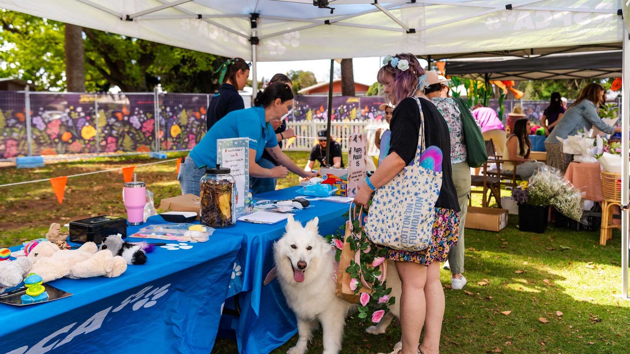 Under a white canopy, a table is covered with a blue cloth displaying various items, including toys and a jar. A person in a blue shirt is interacting with visitors. A fluffy white dog stands beside a person holding a tote bag and a bag with pink flowers. In the background, a colourful mural with floral designs is visible, along with other stalls and people. The ground is grassy with scattered leaves.