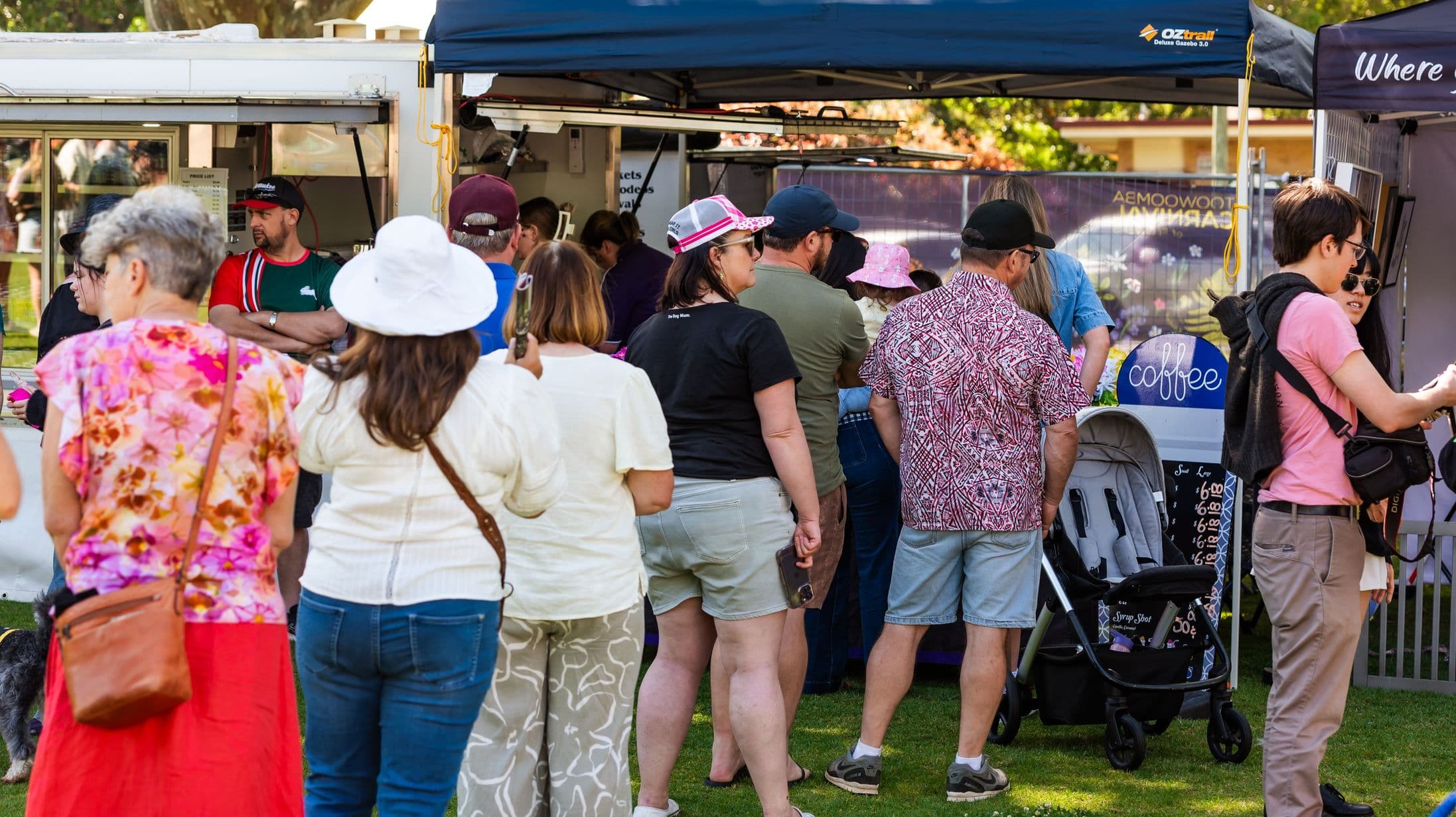 A group of people are queuing in front of a food stall. On the left, a woman in a floral top and red skirt stands next to a woman in a white hat. In the centre, a man in a patterned shirt and a woman in a black T-shirt are visible. On the right, a man in a pink shirt and sunglasses is near a coffee sign and a pram. The background features a canopy and a food truck, with a person in a red and green shirt visible inside. The ground is grassy.