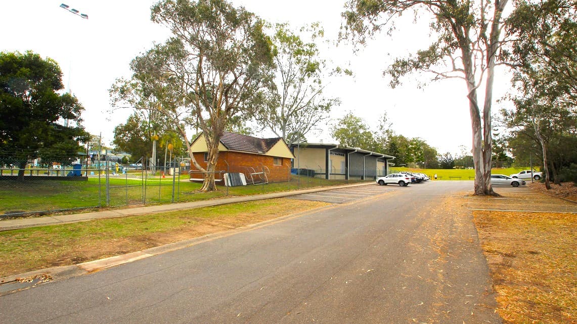 The Ferny Hills Aquatic Centre. Situated amidst suburban residences and nearby schools, there stands a modest brown brick structure that carries a distinct 1980s aesthetic. The presence of mature, expansive trees contributes to a serene ambiance, providing a peaceful atmosphere despite its suburban location. Within the accessible parking spaces, several cars are neatly parked.