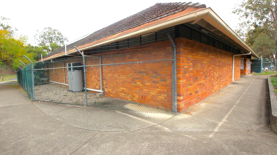 A closer look at the brown brick building. The slender walkway of the building appears to be in good condition and respectable. In the backyard, there are elevated metal poles accompanied by fences. A number of trees are also present in this specific section. The roofing design is reminiscent of the style commonly seen way back in the 80s.