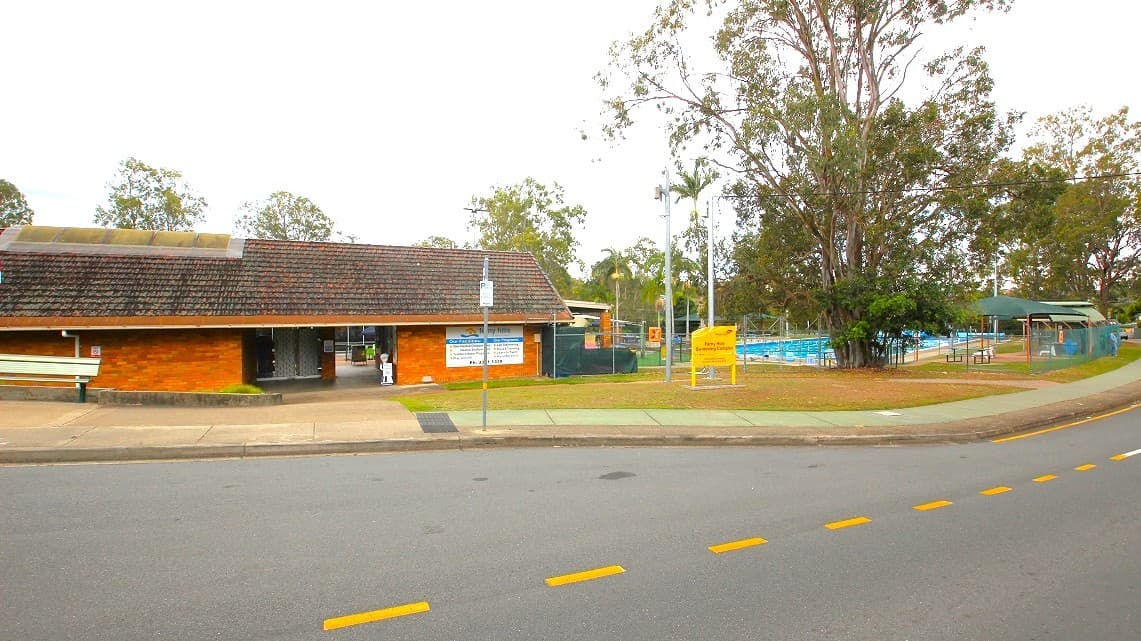 Frontal view of the entrance at Ferny Hills Aquatic Centre. Plenty of gum trees are prominently observable along this side of the road, along with the open-air 50m pool. A set of electric poles with yellow signs underneath them is also present. The distinct 1980s-style brown brick building is an amazing feature.