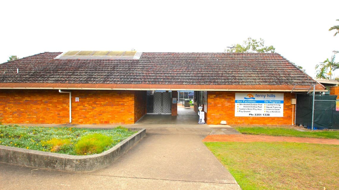 Main entrance leading to Ferny Hills Aquatic Centre. The entrance is not only large but also impeccably neat. The unmistakable brown brick structure, reminiscent of the 1980s style, is easily discernible from this vantage point. Additionally, decorative items have been positioned within a cemented box used as a planter in the garden. The bricks composing the wall appear to have been diligently preserved over the passage of time.
