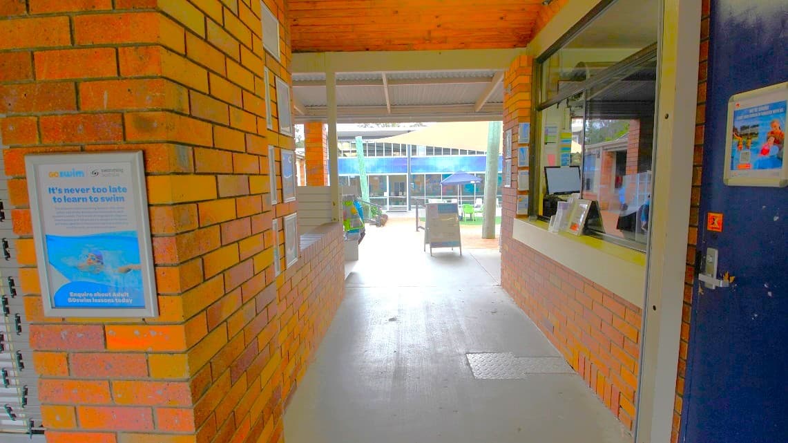 Inside the reception area of Ferny Hills Aquatic Centre. A number of decorative frames are positioned on the left side of the main entrance through the building. Within the brown brick building, a minor passageway is illuminated by fluorescent lights at a low to moderate intensity. The counter of the reception hall is situated on the right-hand side.